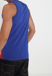 Camisa sin mangas azul de algodón, con un respaldo liso y acentos rojos a los lados, combinada con jeans de mezclilla oscura.