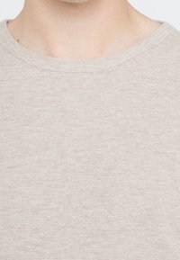 Manlig nacke och övre bröst iklädd en ljusbeige strukturerad stickad t-shirt med rundad hals mot en vit bakgrund.