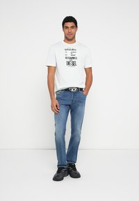 White graphic T-shirt with black text, blue jeans, and black sneakers. The belt features a round silver buckle. Casual fit with a relaxed style.