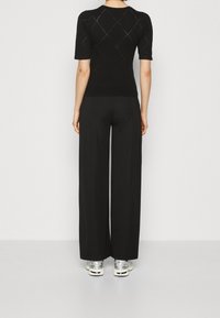 En person som bär en svart kortärmad topp med diamantmönster, vida svarta byxor och silverfärgade sneakers, stående mot en enfärgad vit bakgrund.