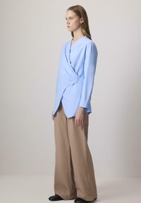 Kék, hosszú ujjú, V nyakú, oldalt megkötős ing, bézs, széles szárú nadrággal párosítva, fekete cipőkkel kiegészítve. Sima textúrájú anyag.