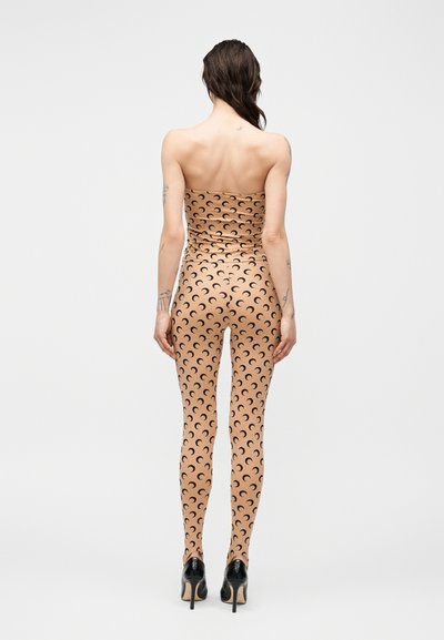 Ženska z dolgimi temnimi lasmi stoji obrnjena stran, oblečena v bež kombinezon brez naramnic s črnimi vzorci polmeseca in črnimi visokimi petami.