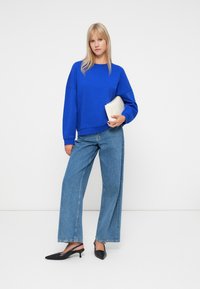 Hanorac oversized albastru, jeansi evazați de culoare albastru deschis, mule cu toc negre și o poșetă albă. Ținuta are un croi lejer și un stil casual.