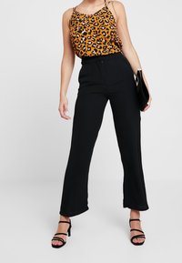 Haut à imprimé léopard orange et noir, associé à un pantalon large noir taille haute. Le mannequin tient une pochette noire et porte des talons à brides.