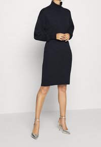 Abito lungo blu navy con maniche lunghe, collo a imbuto finto, silhouette aderente e texture liscia. Indossato con tacchi a punta argentati.