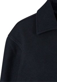 Détail d'épaule et large col d'un manteau en laine noire avec coutures apparentes sur fond blanc.