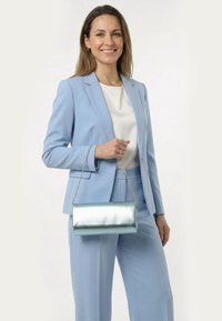 Femme en costume bleu clair tenant un sac à main argenté métallique avec une bandoulière chaîne, souriante sur un fond blanc uni.