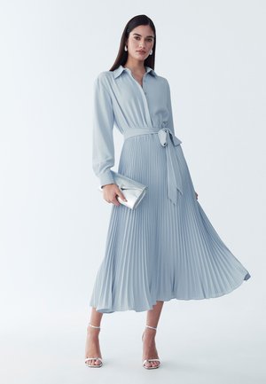 Femme portant une robe midi plissée bleu clair à manches longues, tenant une pochette argentée et portant des sandales argentées à talons hauts.