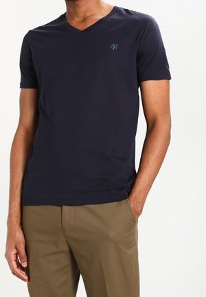 Hombre vistiendo una camiseta de manga corta azul marino con cuello en V y pequeño logo en el pecho, y pantalones caqui, de pie con una mano en el bolsillo.