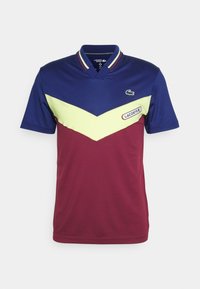 Kortärmad polo t-shirt med en marinblå topp, gul chevrondesign och vinröd botten med strukturerad tyg och Lacoste-logotyp på framsidan.