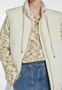 Světle krémová péřová vesta s vysokým límcem a šňůrkami, vrstvená přes texturovaný béžový a olivový pletený svetr, doplněná modrými džínami.