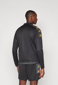 Veste de sport noire à manches longues et col. Accents en logo doré sur les épaules. Tissu lisse et coupe décontractée, accompagnée de shorts assortis.