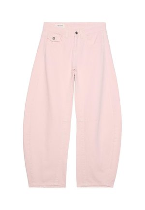 Žena stoji uz teksturirani zid noseći prozirni ružičasti čipkasti top i široke blijedo ružičaste hlače s crnim sandalama, gledajući ulijevo.