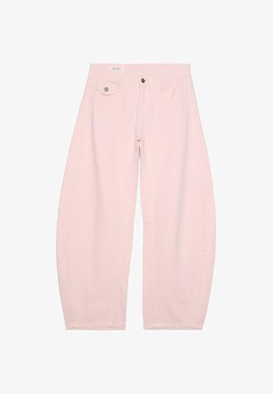 Frau steht vor einer strukturierten Wand, trägt ein transparentes rosa Spitzenoberteil und weite hellrosa Hosen mit schwarzen Sandalen und schaut nach links.