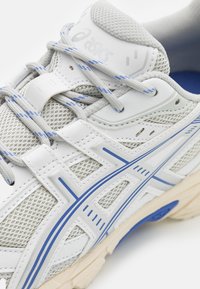 Baskets blanches avec une tige en mesh gris, accents bleus et semelle texturée. Équipées de lacets et de marquages sur la languette et les côtés.