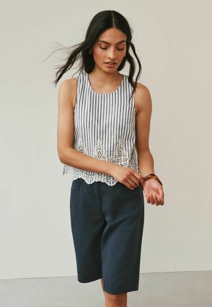 Frau mit langen dunklen Haaren trägt ein ärmelloses gestreiftes Oberteil mit Spitzenabschluss und marineblaue knielange Shorts, während sie ein Armband an ihrem Handgelenk einstellt.