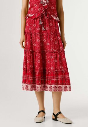 Vrouw draagt een gelaagde rode midi-rok met bloemenprint en kwastdetails, gecombineerd met beige platte schoenen met zwarte enkelbandjes, staand tegen een witte achtergrond.