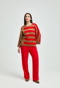 Brauner Strickpullover mit roten Streifen, kombiniert mit roten weiten Hosen. Weiche Textur, lockere Passform, runder Ausschnitt und minimalistisches Design.