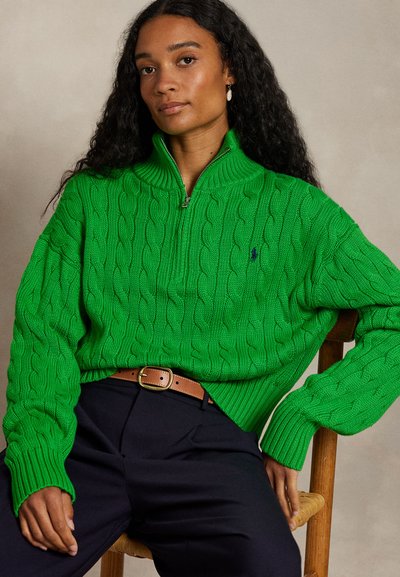 Πράσινο πουλόβερ με πλέξη καλτσόδερου, ψηλό γιακά και κλείσιμο με φερμουάρ στη μέση. Διαθέτει ριμπ μανσέτες και μικρό λογότυπο στο στήθος. Φοράει πάνω από σκούρο παντελόνι.