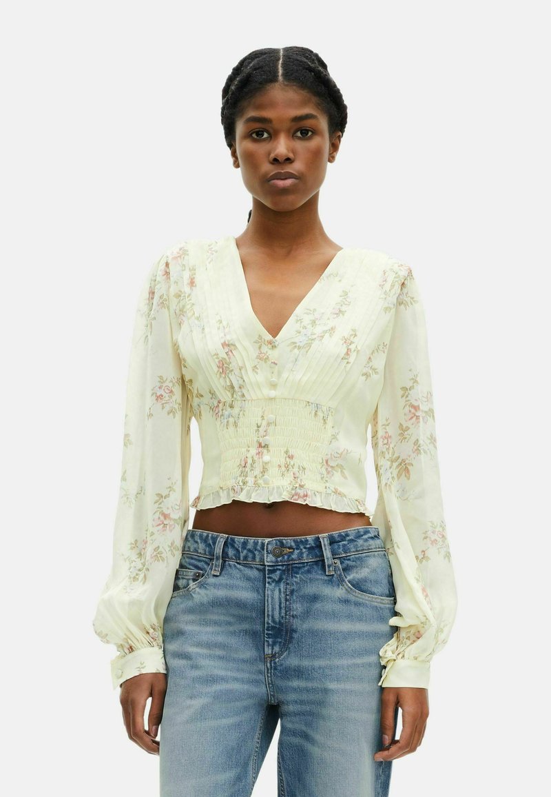 Top floreale cropped in morbido tessuto giallo con dettagli plissettati, scollatura a v e maniche lunghe a palloncino; abbinato a jeans in denim chiaro.