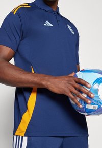 Homme en tenue de sport bleue et jaune tenant un ballon de football bleu et blanc sur un fond uni.