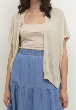 Kobieta ubrana w kremowy dziergany bezrękawnik z pasującym krótkim sweterkiem na ramiona oraz niebieską dżinsową spódnicę z wysokim stanem i elastycznym paskiem.