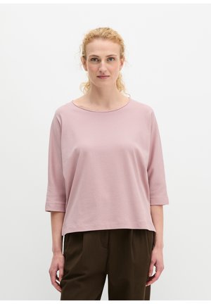 Femme aux cheveux blonds bouclés porte un haut rose clair ample à manches trois-quarts et un pantalon marron foncé, debout devant un fond blanc uni.