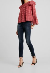 Rød blomstret blouse med flæser på ærmerne, parret med mørke skinny jeans og sorte højhælede sandaler, som fremviser et lagdelt design og tekstur.