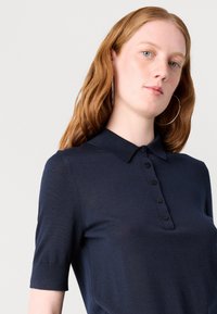 Tricou polo de culoare bleumarin, confecționat dintr-un material moale, având guler, mâneci scurte și un închidere cu nasturi, dotat cu șase nasturi negri.