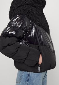 Svart puffjacka med en blank yta på ärmarna och ett texturerat, fluffigt tyg på överkroppen. Dragkedjedetaljer ingår.