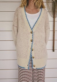 Džemper kardigan u krem boji s uzorkom točkica, plavim naboranim rubom duž ivica i smeđim gumbima. Uparen sa prugastim hlačama.