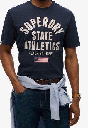 Mężczyzna ubrany w granatowy t-shirt Superdry State Athletics, niebieskie dżinsy, jasnoniebieską koszulę zawiązaną wokół talii oraz brązowy zegarek na lewej ręce.