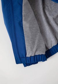 Modrá větrovka s elastickými manžetami; šedá podšívka; lehký materiál s hladkou texturou; jednoduchý a funkční design.