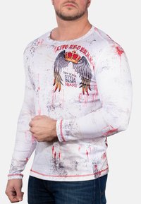 Langärmliges weißes Hemd mit einem abgenutzten Textur, verziert mit roten Farbklecksen, einem Flugmotiven-Druck und einem Kronendesign auf der Brust.
