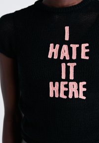Suéter negro de punto de manga corta con gran texto bordado en rosa que dice "ODIO ESTAR AQUÍ" centrado en la parte frontal.