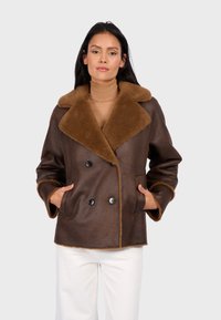 Brązowy płaszcz z owczej skóry z dużym brązowym kołnierzem, podwójnym zapięciem na guziki, czarnymi guzikami i kontrastowymi jasnobrązowymi wykończeniami. Model pozuje z rękami w kieszeniach.