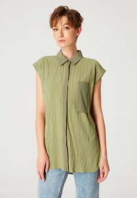Chemise vert olive à manches courtes avec texture de crêpe verticale, bouton sur le devant, col pointu et poche sur la poitrine gauche. Portée avec un jean bleu clair.