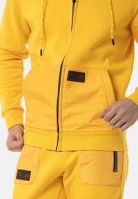 Gelbes Hoodie mit Frontreißverschluss, gerippten Bündchen und Taschen, mit einem Stoffpatch-Label. Material hat eine glatte Textur.