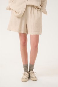 Beige, gestructureerde shorts met een elastische tailleband, gecombineerd met grijze, geribde sokken en lichte sneakers met een subtiel patroon.