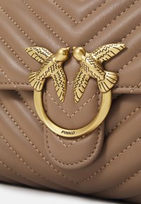 Viereckige Handtasche aus weichem beigen Leder mit gestepptem Zickzackmuster. Verfügt über einen goldfarbenen rundenverschluss mit zwei Vogelakzenten.