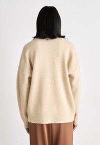 Beige oversized sweater with a soft, textured finish, drop shoulders, and ribbed hem, featuring a small black logo at the upper back.