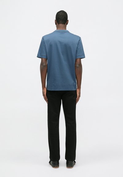 T-shirt de algodão azul com mangas curtas, gola redonda e um corte relaxado, combinada com calças pretas de perna reta e sapatos pretos, vista de costas.