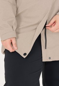 Beiger Jackenärmel und Saum mit Druckknopf und Reißverschluss, gehalten von Händen, kombiniert mit schwarzer Hose.