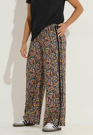 Osoba nosząca spodnie z szerokimi nogawkami w wielokolorowy kwiatowy wzór z czarnymi bocznymi paskami, do tego czarny top i białe trampki z czarnymi paskami.