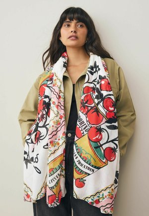 Femme avec de longs cheveux bruns portant une veste beige et une écharpe colorée avec des tomates rouges et un texte sur un top noir et un pantalon sombre.