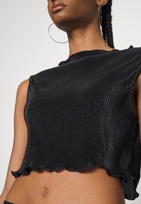Femme portant un crop top sans manches côtelé noir et de grandes boucles d'oreilles en anneaux, montrant le cou, les épaules et le ventre sur un fond uni.