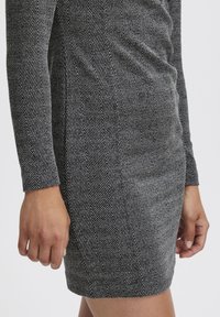Črna in bela obleka s herringbone vzorcem z dolgimi rokavi, narejena iz teksturirane tkanine. Rob je nekoliko nad kolenom.
