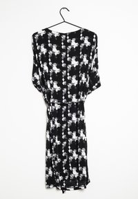 Svart blommig klänning i lätt tyg, med ett geometriskt vitt mönster, korta ärmar och en lös passform, hängande på en svart krok.