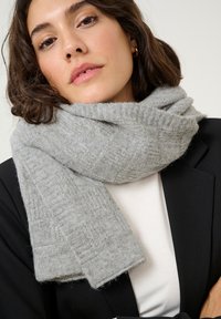 Écharpe tricotée gris clair avec un motif texturé, enroulée autour du cou. Portée avec un blazer noir sur un haut blanc.