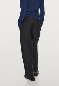 Donkergrijze broek met een wijde pijp, zijzakken en knoopzakken aan de achterkant. Draagt een marineblauwe trui en donkere schoenen.